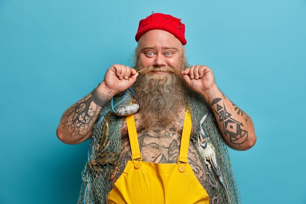 Glad fisherman curls mustache, has thick beard, carries fishing net on shoulders, spends free time for hobby and soul, wears red hat and overalls, has tattoed body