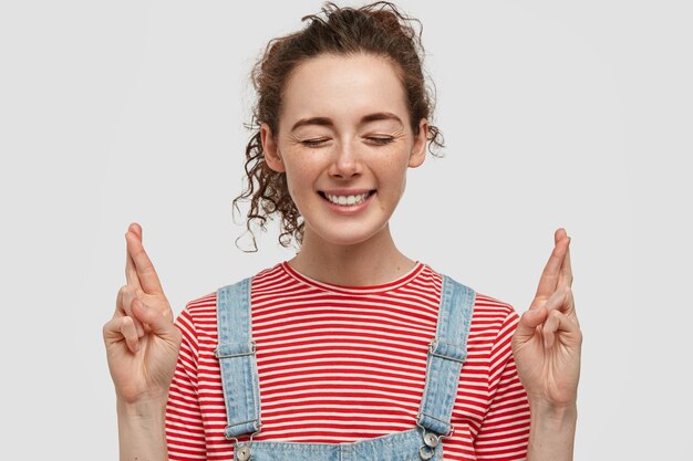 Glad freckled young female closes eyes, has broad smile, crosses fingers as makes wish, dressed in pink striped t-shirt and dungarees, stands against white wall. Woman prays for better.