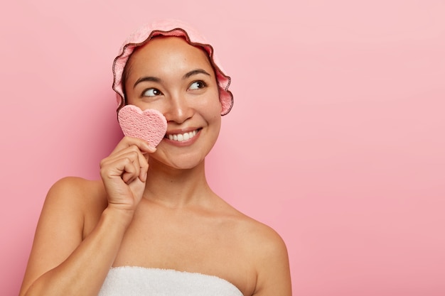Glad fresh young woman cleans face with sponge, has dreamy happy expression, wears bathcap, recalls pleasant moment in memory