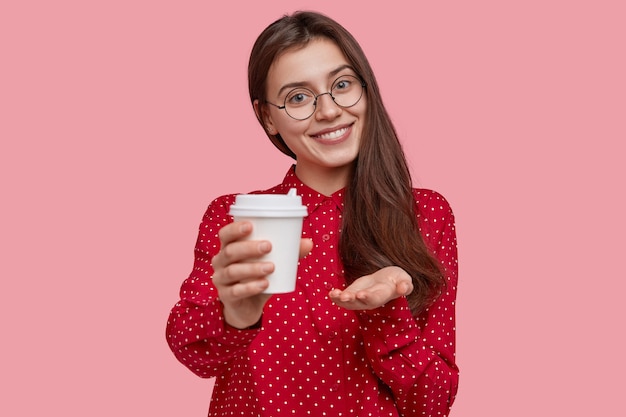 Glad friendly looking woman holds disposable cup of coffee, suggests to drink together, wears optical glasses, red polka dot blouse, tilts head