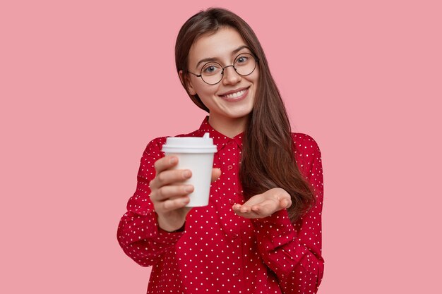 Glad friendly looking woman holds disposable cup of coffee, suggests to drink together, wears optical glasses, red polka dot blouse, tilts head