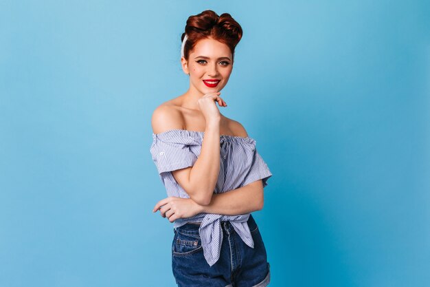 Glad ginger girl looking at camera with smile. Studio shot of attractive pinup female model isolated on blue space.