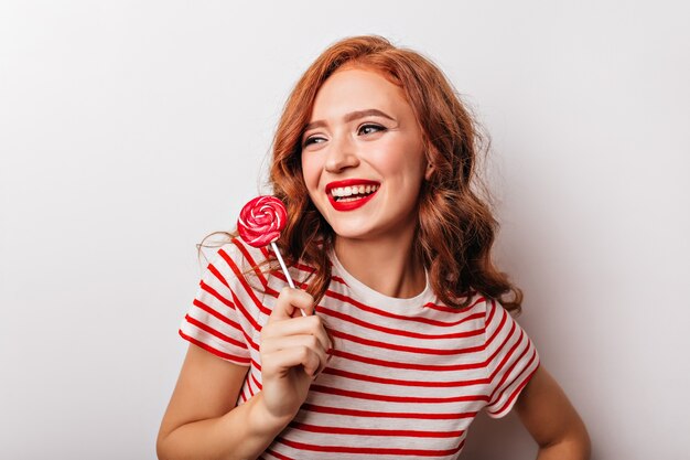 Glad ginger woman holding lollipop and laughing. Glamorous caucasian girl with candy enjoying photoshoot.