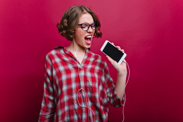 Glad girl in glasses singing favorite song on claret wall. Indoor portrait of wonderful short-haired young woman enjoying music and holding phone.