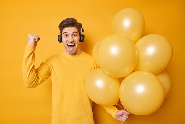 Glad handsome man celebrates successfully passed exams being on leaving party listens music via headphones holds bunch of yellow helium balloons poses indoor wears casual jumper Celebration happiness