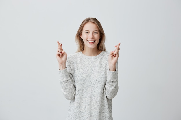 glad happy blonde woman wears sweater, smiles broadly, keeps fingers crossed, hopes for good luck