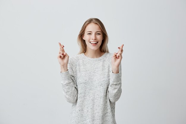 glad happy blonde woman wears sweater, smiles broadly, keeps fingers crossed, hopes for good luck