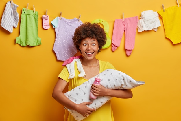 Free photo glad happy mum embraces her little infant, holds bottle with nipple and feeds baby, nursing newborn, prepares artifical feed, stands against yellow wall, washed childs clothing hanging on rope