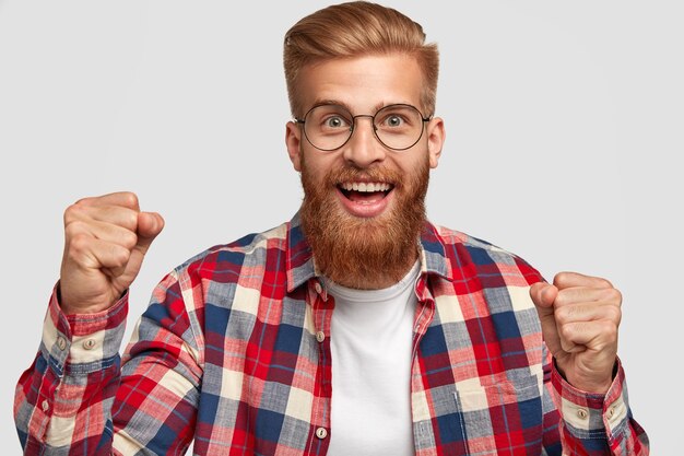 Glad hipster with funny expression, clenches fists, celebrates successful day, has trendy hairstyle and ginger beard, wears bright checkered shirt, isolated over white wall. Triumph concept