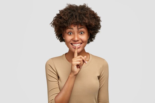 Glad lovely African American female asks to keep silence, holds index finger on lips, has cheerful expression, dressed casually, poses against white wall. People, secrecy and ethnicity concept