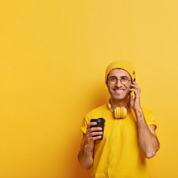 Glad male model calls friend via cellphone, holds mobile phone, enjoys conversation while drinking takeout coffee, wears yellow clothes, transparent glasses