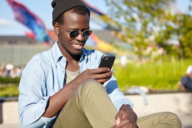 어두운 피부를 가진 다행 남성, 선글라스와 최신 유행의 옷을 입고, 휴대 전화로 즐거운 SMS를 읽고 답을 입력하십시오. 항상 피부에 닿으면서 외부의 스마트 폰을 사용하여 어두운 피부를 가진 남성 미소