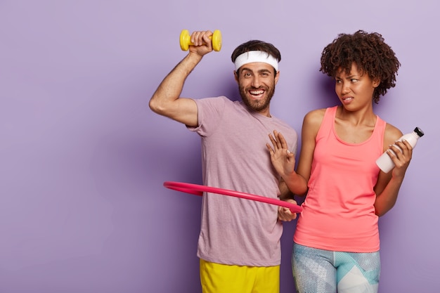 Glad motivated man with stubble, rotates hula hoop, trains muscles with dumbbell and dissatisfied afro woman makes refusal gesture, holds bottle of water