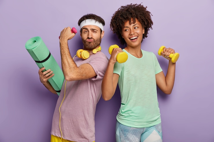 Free Photo glad multiethnic husband and wife attend sport center, exercise with dumbbells, hold fitness mat, stand back to each other, have funny happy looks, wear t shirts, isolated on purple wall