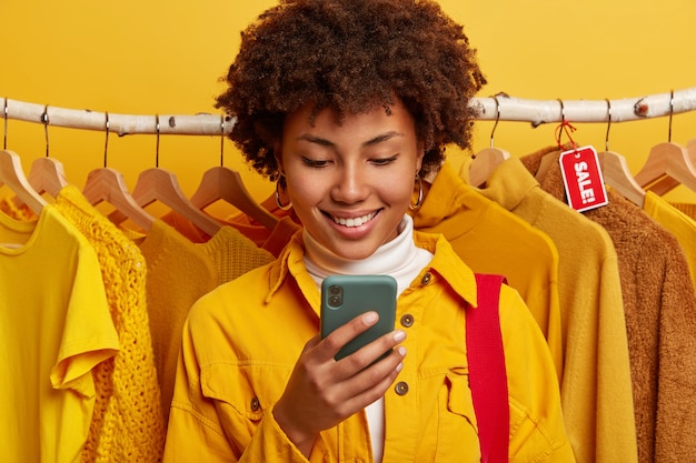 Glad online merchant focused in smartphone device, stands against yellow clothing on racks