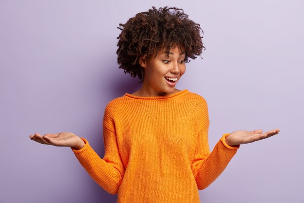 Glad optimistic Afro American woman raises both palms, pretends holding two items, being in good mood, wears casual outfit, isolated over purple wall. Satisfied female holds copy space