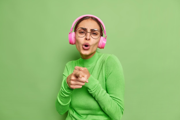 嬉しい遊び心のある女性が歌を歌うことは、ピンクのヘッドフォンでお気に入りの音楽を楽しんでいることを示しています。透明なメガネをかけ、緑の壁にタートルネックのポーズをとっています。
