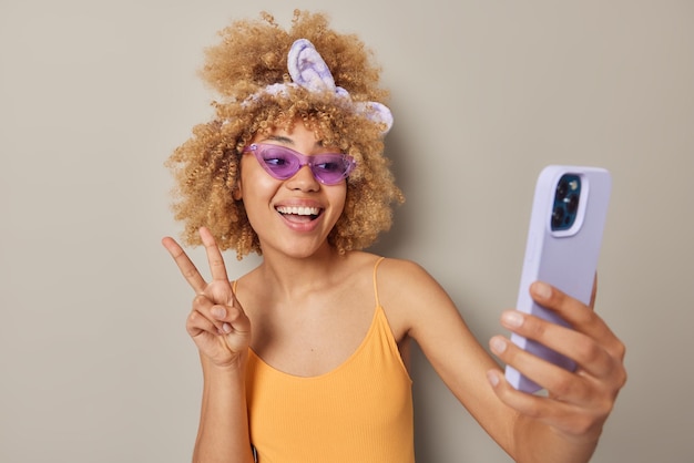Бесплатное фото Рад, что приятно выглядящая европейская женщина с вьющимися волосами носит фиолетовые солнцезащитные очки с повязкой на голове и повседневную футболку делает мирный жест, делает селфи через смартфон, изолированный на сером фоне студии