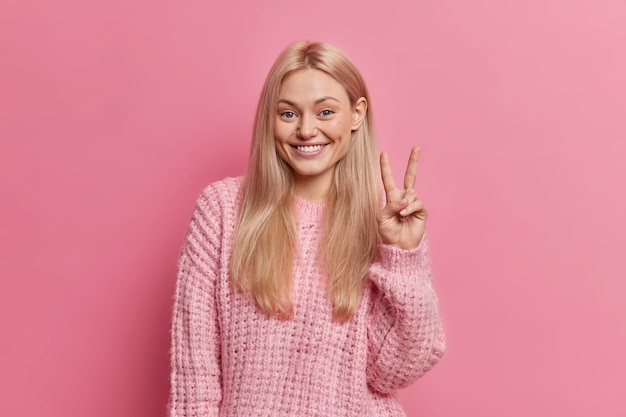 Glad pleasant looking woman with light hair makes peace sign, smiles toothily