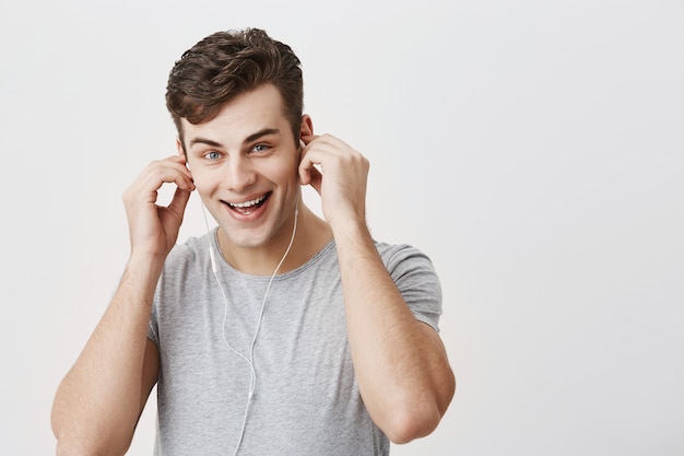 Free photo glad positive caucasian man smiles joyfully, listens music in earphones, holds hands behind his ears, enjoys favourite songs, uses music app. young european male enjoys pleasant melodies alone