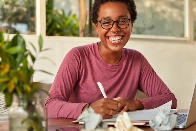 Free photo glad positive dark skinned young female rewrites on blank sheet of paper some information from web page