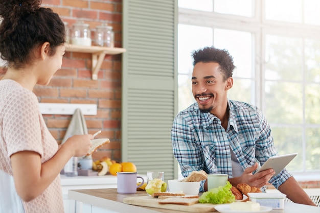 Glad positive hipster guy dressed casually, sits at table, waits for lunch prepared by housewife, holds digital tablet,