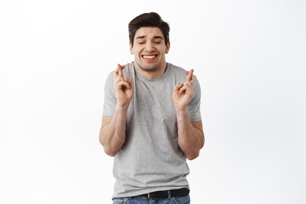 Glad positive man crosses fingers closes eyes with pleasure anticipate hearing good news isolated white background Happy male rejoices that his dreams came true Excitement concept