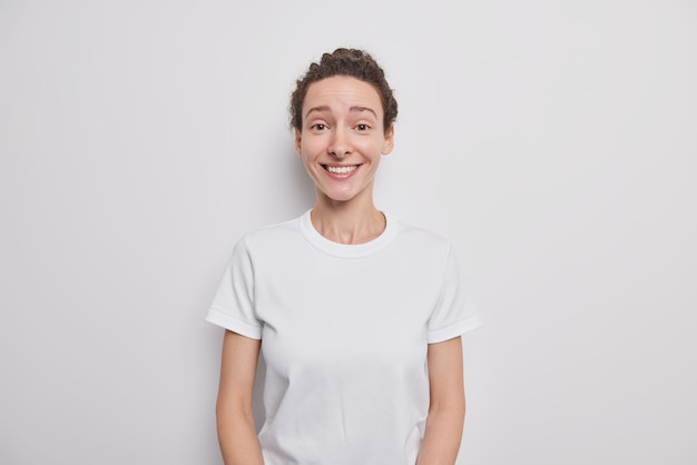 Glad pretty millennial girl with natural beauty smiles happily has dark combed hair dressed in casual t shirt models indoor on white expresses sincere feelings listens smth interesting