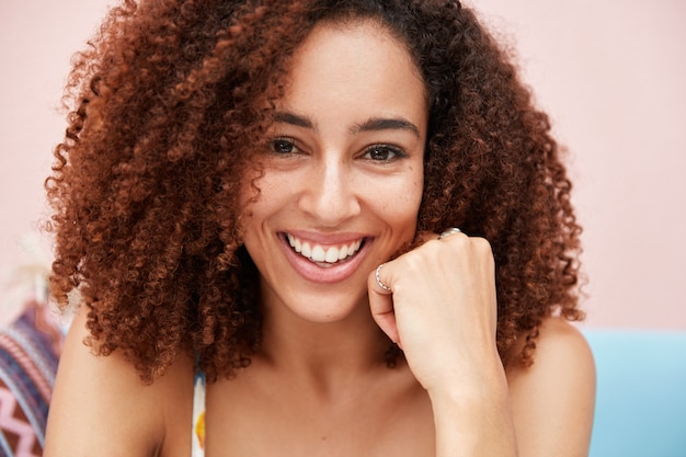 Довольная молодая афроамериканка-модель с широкой нежной улыбкой, с вьющимися вьющимися густыми волосами, смуглой кожей, позирует перед камерой с веселым выражением лица.
