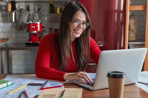 Glad professional brunette female accountant makes distant work, keyboards on laptop computer, sits at kitchen table with papers, wears transparent glasses for good vision, drinks coffee to go