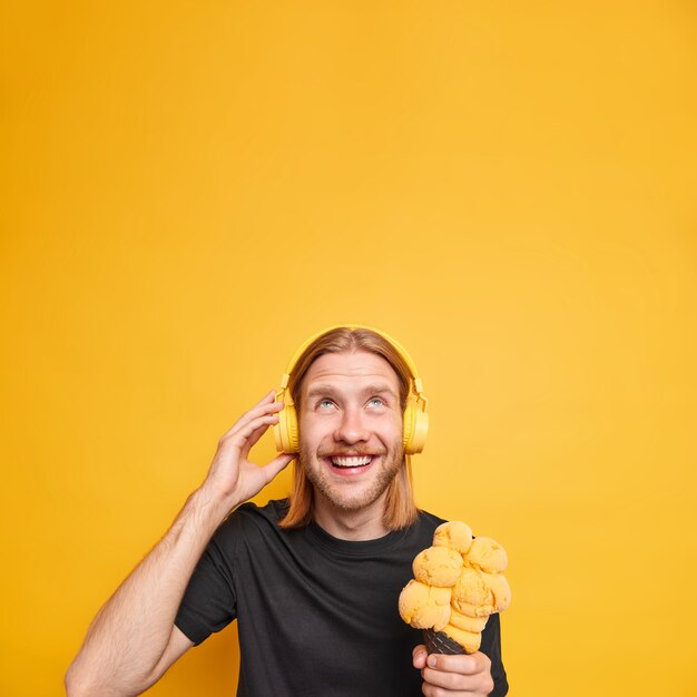 幸せなリラックスした生姜の男が頭上で微笑み、おいしいアイスクリームを広く持ち、ヘッドフォンで音楽を聴く 鮮やかな黄色の壁のコピースペースの上に黒いTシャツを着ている