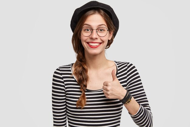 Glad satisfied French woman with attractive appearance, raises thumb, shows her like and agreement