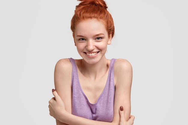 Glad shy female model has freckles on face, red hair combed in knot, dressed in casual oversized clothes