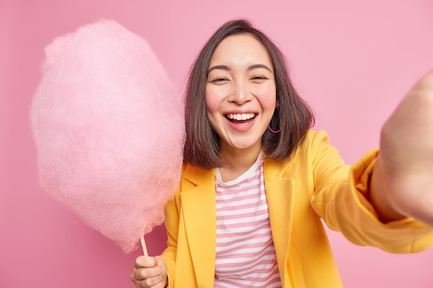 기쁜 성실한 아시아 여성은 정통 감정을 표현하고 맛있는 사탕 치실을 보유하고 있으며 여름 산책 중에 셀카 미소가 긍정적으로 좋은 분위기를 가지고 분홍색 벽에 고립 된 세련된 옷을 입습니다.