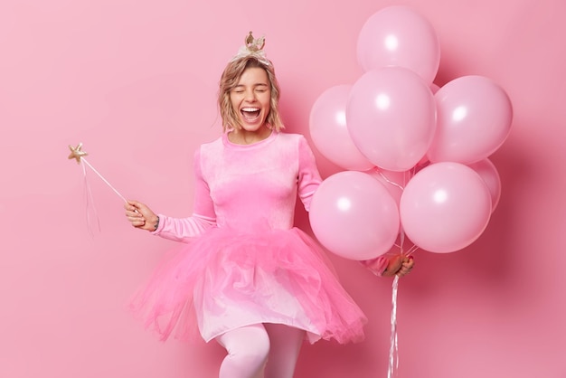 Glad sincere young woman with hairstyle exclaims from happiness wears crown and dress holds magic wand bunch of inflated balloons enjoys celebration and party time isolated over pink background.