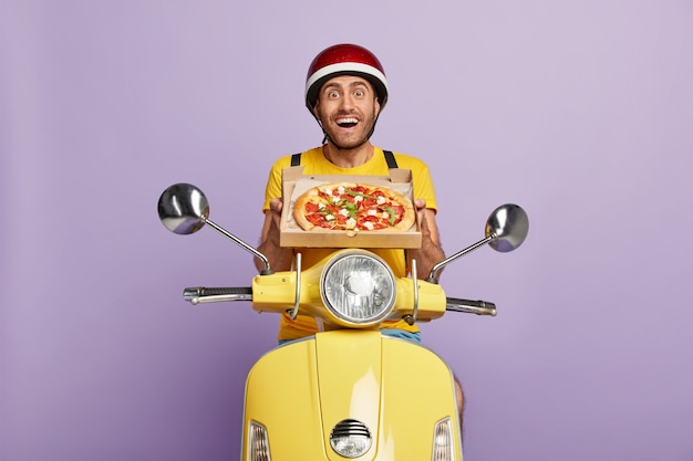 Glad skilled deliveryman driving yellow scooter while holding pizza box