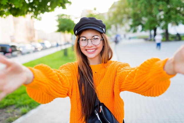 Glad smiling brunette girl in round eyeglasses and trendy orange knitted sweater making self portrait