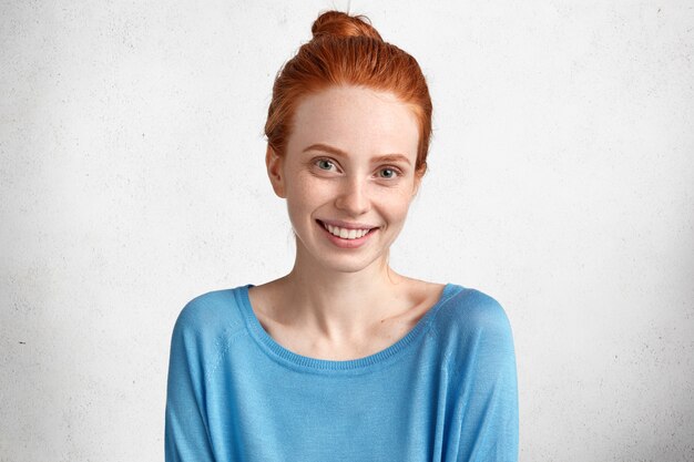 Glad smiling female with freckled skin and positive smile, dressed in blue casual sweater, being in high spirit after date with boyfriend, isolated over white