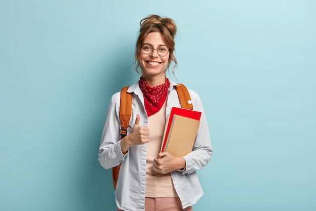 Free photo glad student gives approval, keeps thumb up, holds notebook and diary, has happy smile, says okay, wears optical spectacles