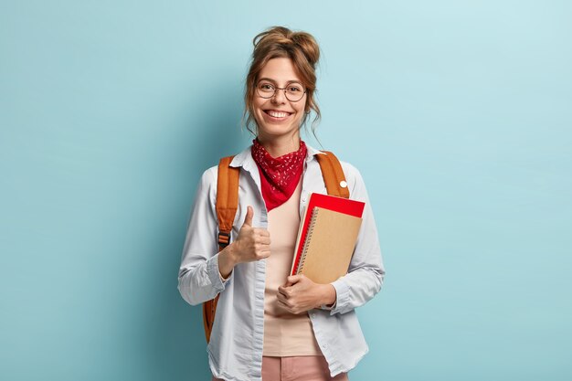 Glad student gives approval, keeps thumb up, holds notebook and diary, has happy smile, says okay, wears optical spectacles