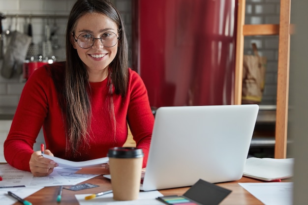 Foto gratuita lo studente felice si prepara per il seminario, scrive i compiti, controlla la documentazione del testo, usa il computer portatile, si siede al tavolo della cucina