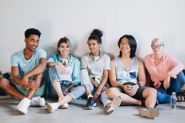 Glad students wearing stylish sneakers and accessories sitting together on the floor with legs crossed. Excited young people of different nationalities relaxing in light room and laughing..