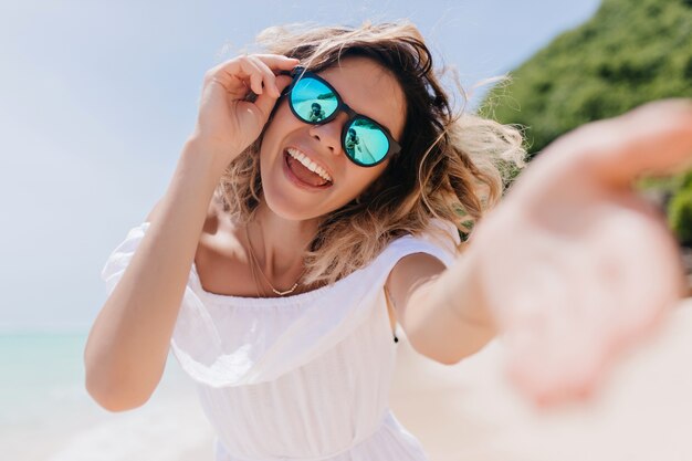 熱帯の島で楽しんでいる輝きのメガネでうれしいスタイリッシュな女性。夏休みの前向きな感情を表現するウェーブのかかった髪の素晴らしい女性の屋外写真。