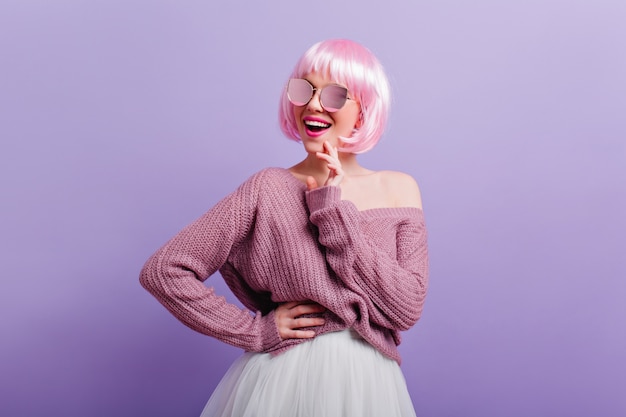 Радостная стильная женщина с розовыми волосами, счастливый смех Крытый портрет улыбающейся экстатической девушки, стоящей в уверенной позе на фиолетовой стене.