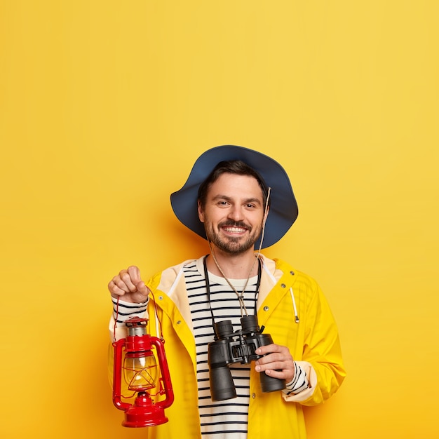 Free photo glad unshaven man carries binoculars and oil lamp, wears hat and raincoat, has mustache, smiles pleasantly, stands against yellow wall
