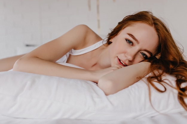 Glad white girl with dark makeup lying on pillow. Sleepy female model with ginger hair posing in bed.