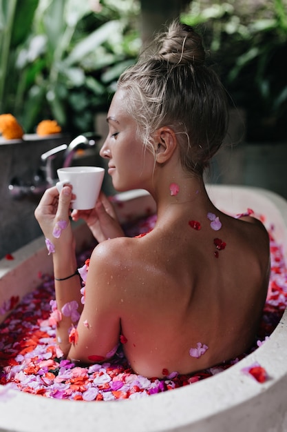 Glad white woman sitting in bath with rose petals and drinking tea with eyes closed. Portrait from back of inspired caucasian female model enjoying coffee during morning spa.