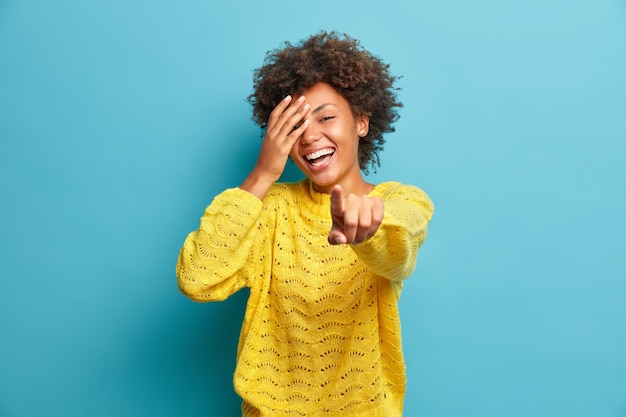 Glad woman cannot hide true happiness points directly at camera expresses good mood dressed in casual sweater expresses sincere emotions sees something funny in front isolated on blue