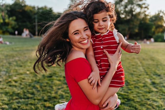 Felice donna che tiene la figlia e che ride alla telecamera. foto all'aperto di giovane mamma emotiva rilassante nel fine settimana con il bambino.