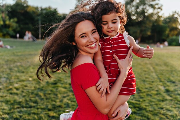 Glad woman holding her daughter and laughing to camera. Outdoor photo of emotional young mom relaxing in weekend with kid.